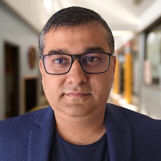 Dipender Bhamrah, head and shoulders, wearing glasses and a dark blue blazer, in front of a blurred corridor.