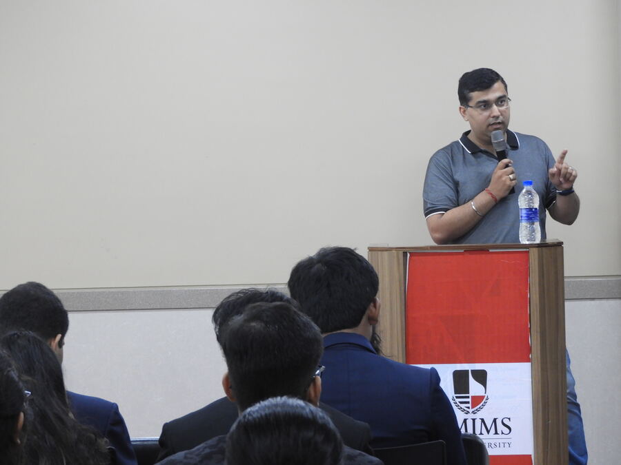Dipender Bhamrah speaking at a lectern in front of a seated student audience at NMIMS, Navi Mumbai